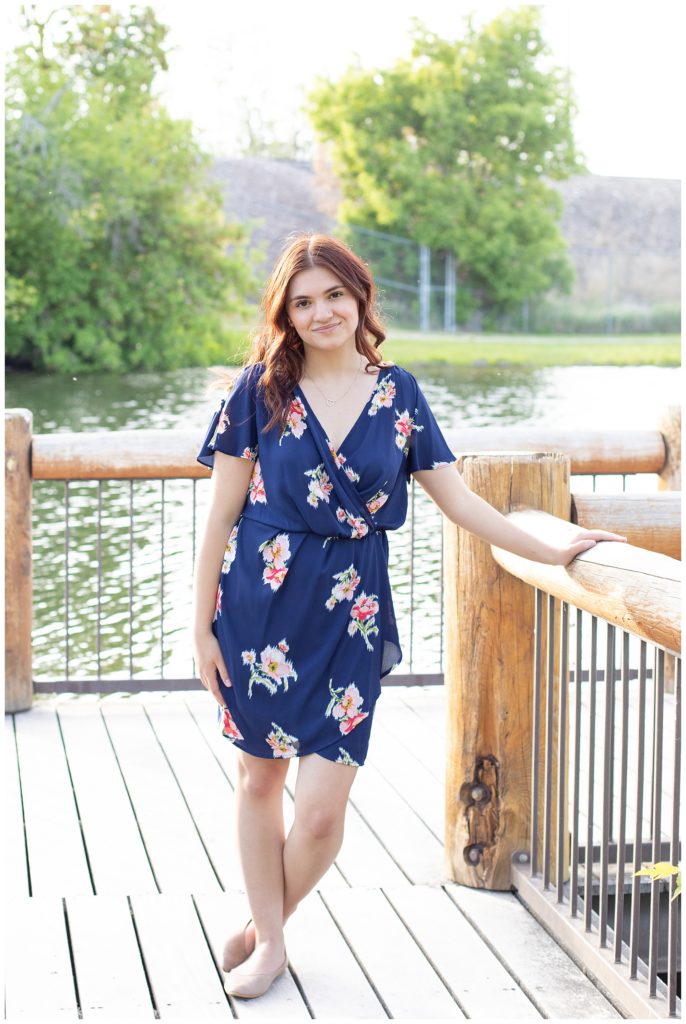 Girl wearing a navy blue dress with pink flowers leans against railing on dock at Silver's Lagoon in Missoula