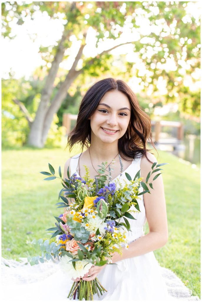Senior girl wearing white sundress sits in grass smiling holding a bouquet of pastel flowers
