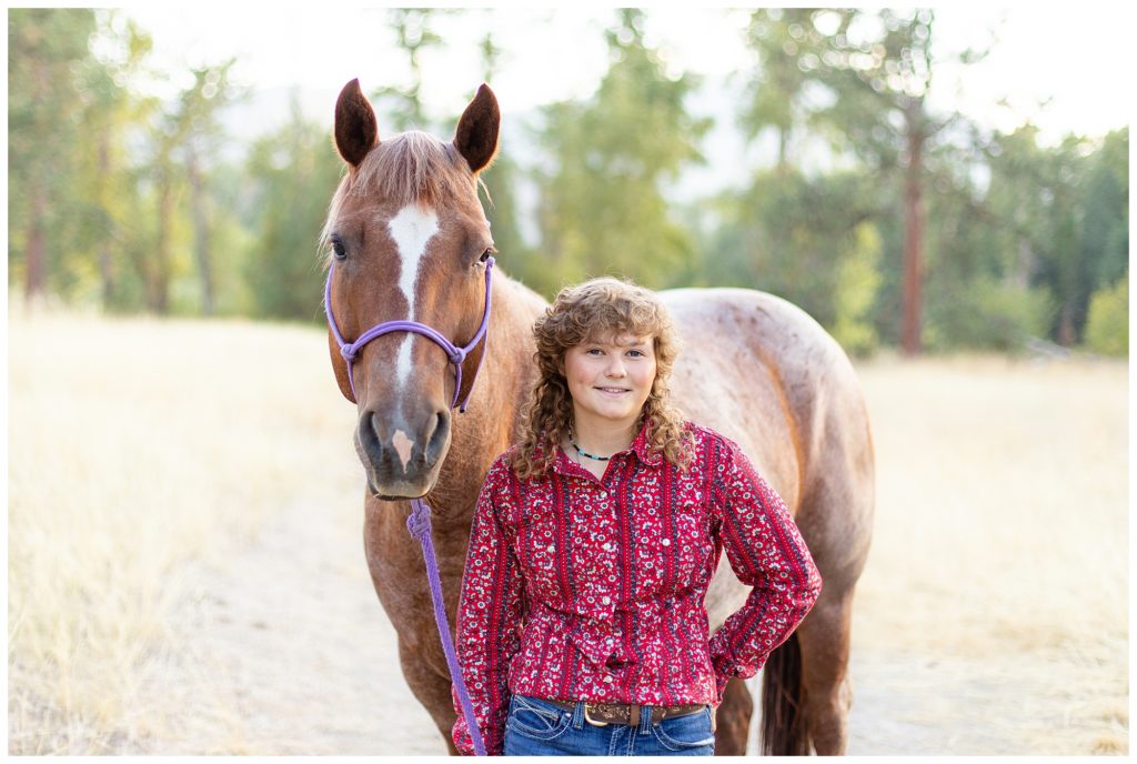 Girl wearing red shirt and jeans smiles next to her roan horse in a field