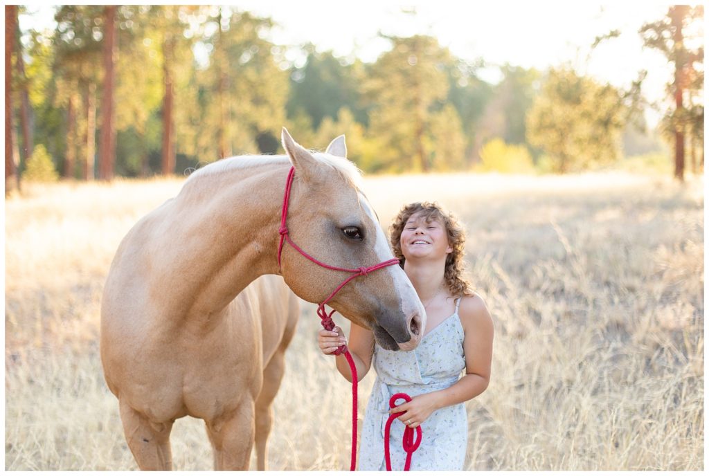 High school senior wearing blue sundress laughs next to her palamino gelding in a field of grass at Council Grove State Park