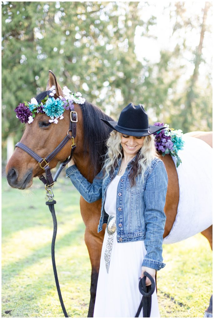 Woman wearing white dress and denim jacket standing next to her horse wearing black leather halter and florals 