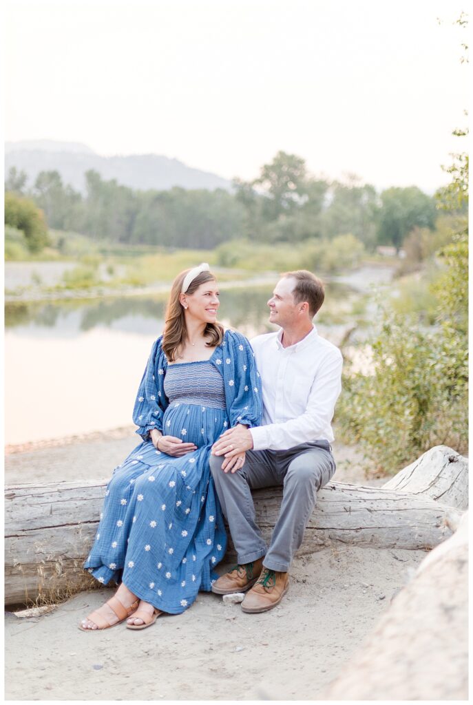 Missoula maternity photos of wife wearing blue dress with flowers and husband in white shirt and gray pants smiling on beach